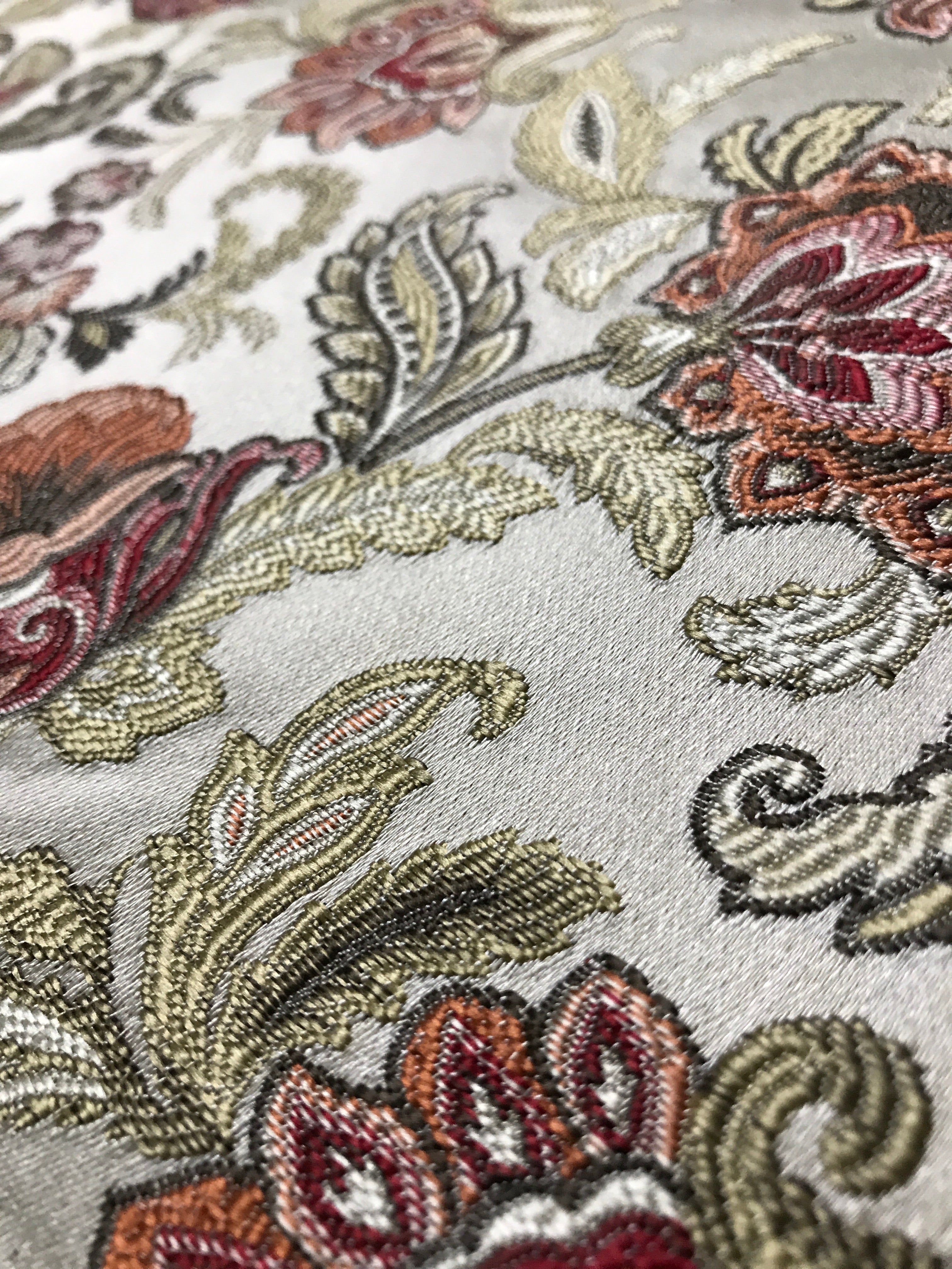 Close-up of embroidered fabric with floral and paisley patterns in a Sage Rose color on a beige background